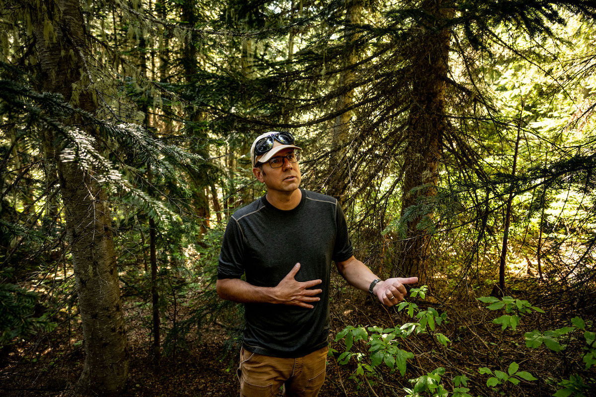 A man with light skin tone wears a T-shirt, duck canvas pants and a baseball cap. He’s standing in the forest.