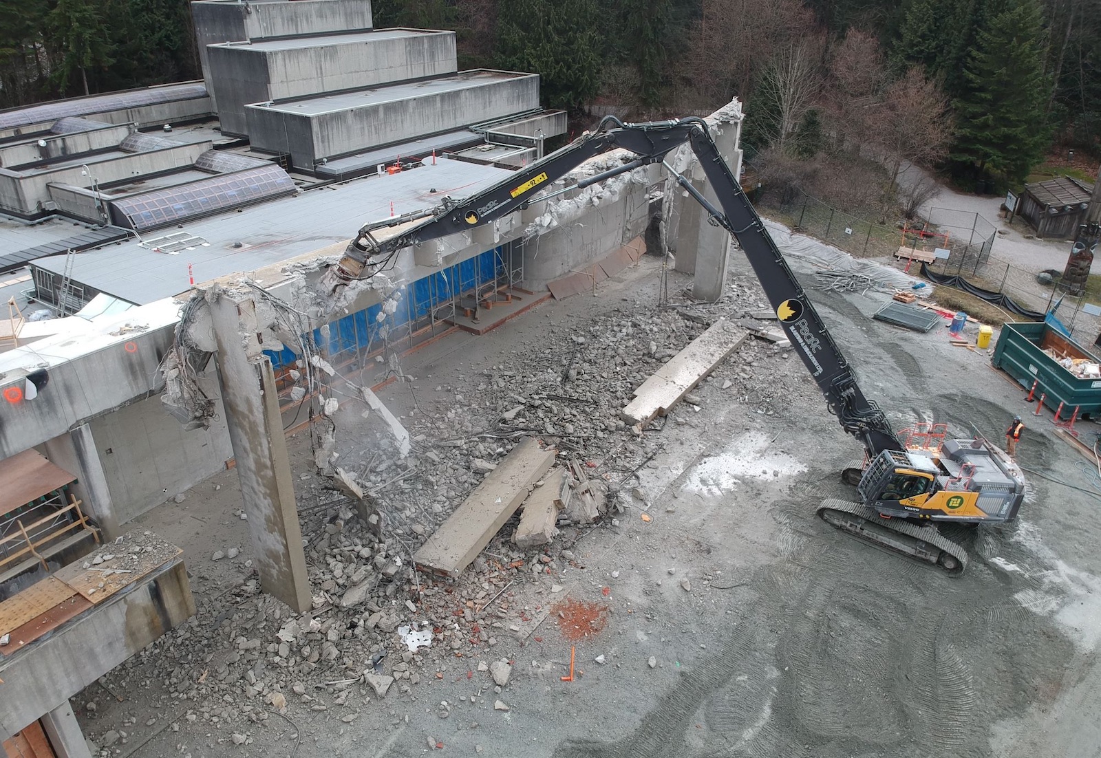 Aerial view of a concrete building with part of it demolished. Concrete rubble and an excavator can be seen in front of the building.