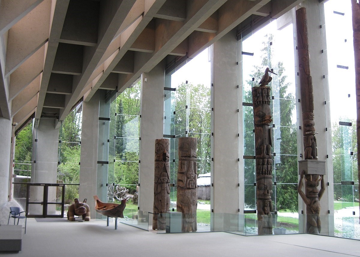A museum hall constructed of floor-to-ceiling glass panes and bare concrete beams houses four wooden totem poles, a small wooden canoe and a wooden animal sculpture. Trees and greenery can be seen through the glass panes.