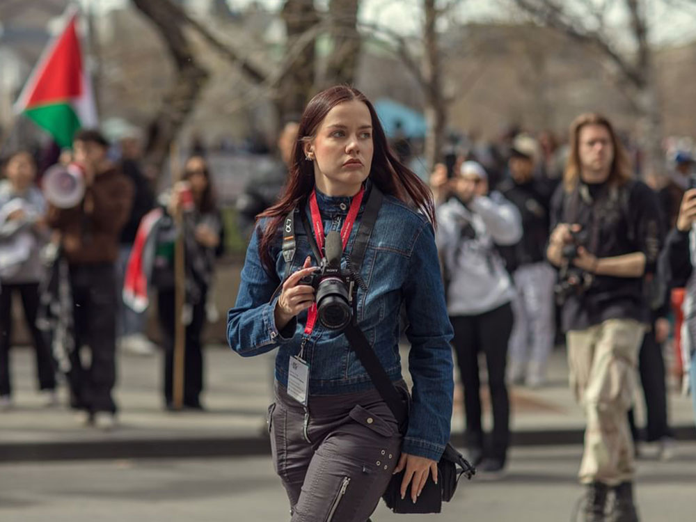 A young woman with light skin and long dark purple hair wears a blue jean jacket and grey pants. She holds a camera around her neck and is looking to the right of the frame. Behind her are people participating in a pro-Palestinian rally.