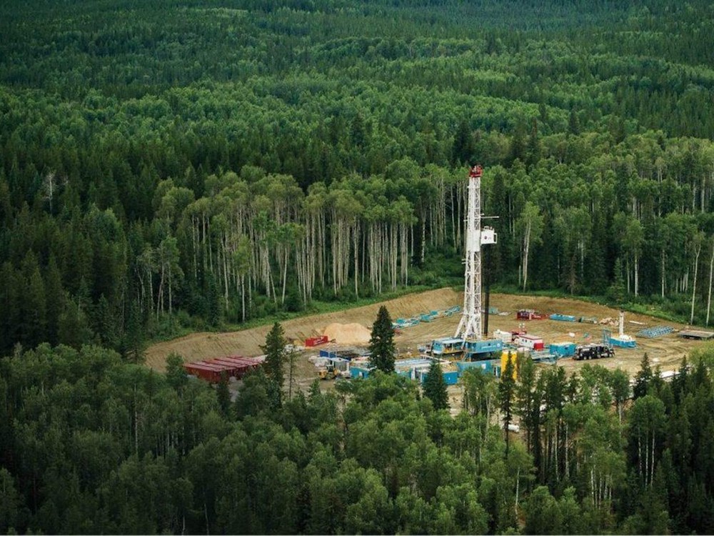 A square oil drilling pad is carved out of the surrounding lush green forest. A white oil tower stands in the centre of the pad, surrounded by containers and machinery.