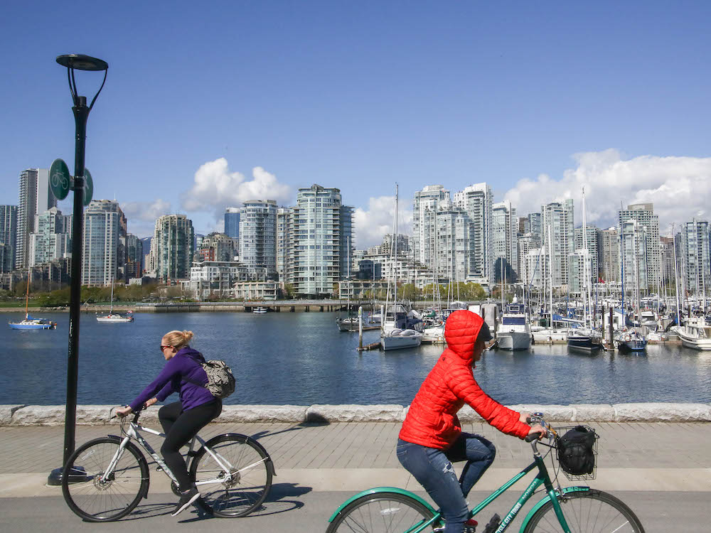 A view from the Vancouver seawall on a sunny winter day. Across the water are glassy highrise towers. In the foreground are two cyclists biking in opposite directions. The cyclist on the left is wearing a dark purple jacket and has blond hair. The cyclist on the right is wearing a bright red puffer jacket with the hood pulled up. The sky is clear and blue.