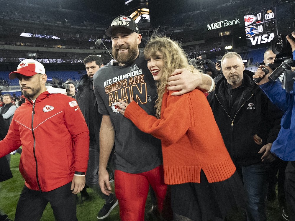 A large, bearded white man in a black ball cap and football jersey stands with his arm around a woman with long blond hair, an orange sweater and black skirt.