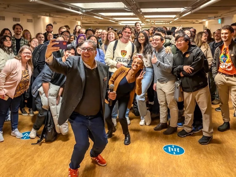 A middle-aged man with medium-dark skin is taking a selfie in front of a group of smiling people. He wears red sneakers, jeans, a brown sweater and a blazer.