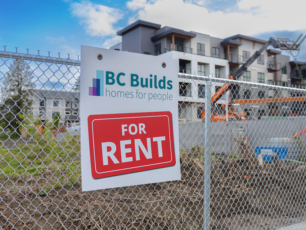 A chain-link fence in front of a vacant lot carries a sign that says 'BC Builds, homes for people, For Rent.' An apartment building is in the background.