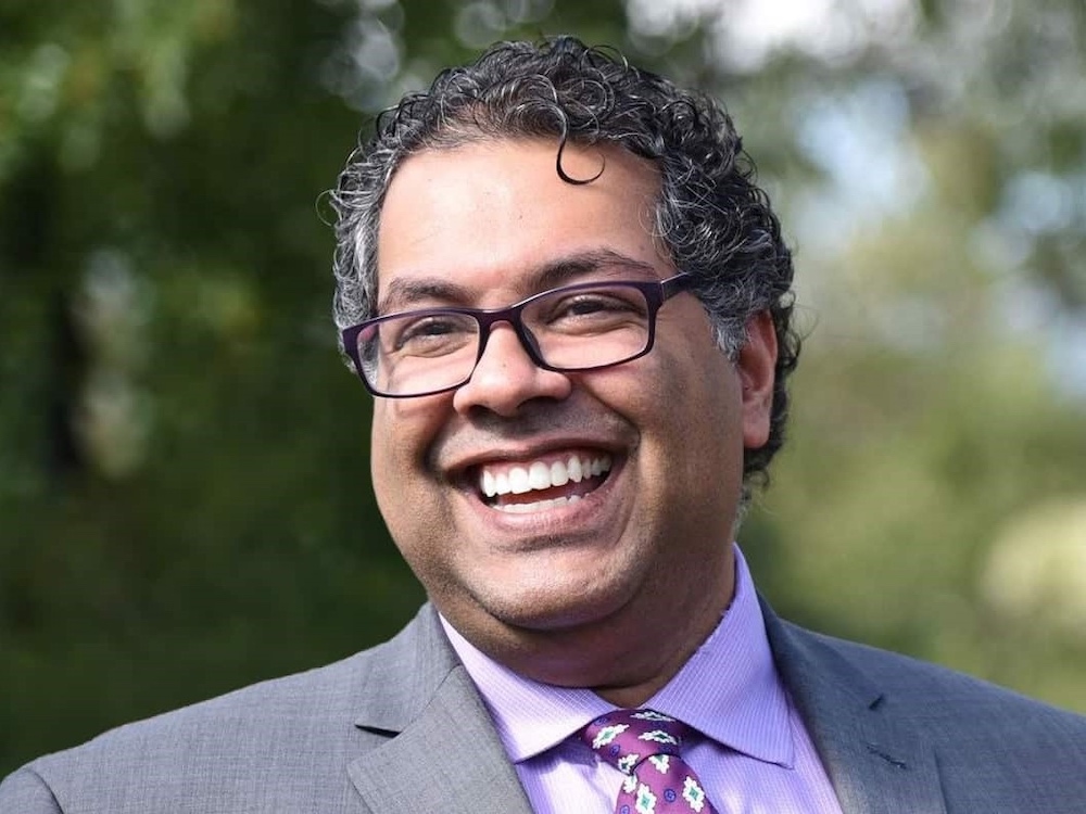 A 52-year old man with medium-toned skin smiles, showing white teeth. He has short curly greying hair and wears a light-grey suit, lilac shirt and purple patterned tie and glasses.