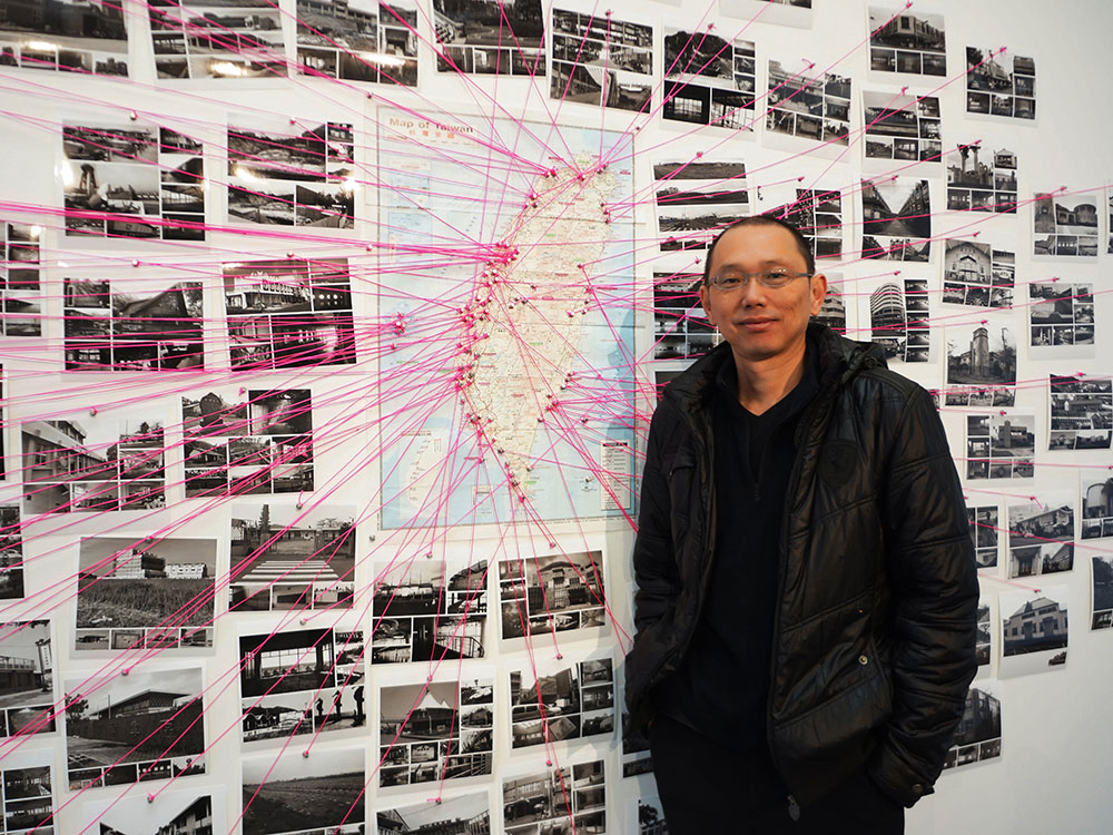 A man w/ medium-coloured skin and buzzed black hair stands in front of a wall of black-and-white photographs and a map of Taiwan. Points on the map are connected the various photos with bright pink string.