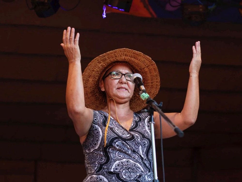 A middle-aged woman wearing a woven hat and glasses raises her arms in front of a microphone.