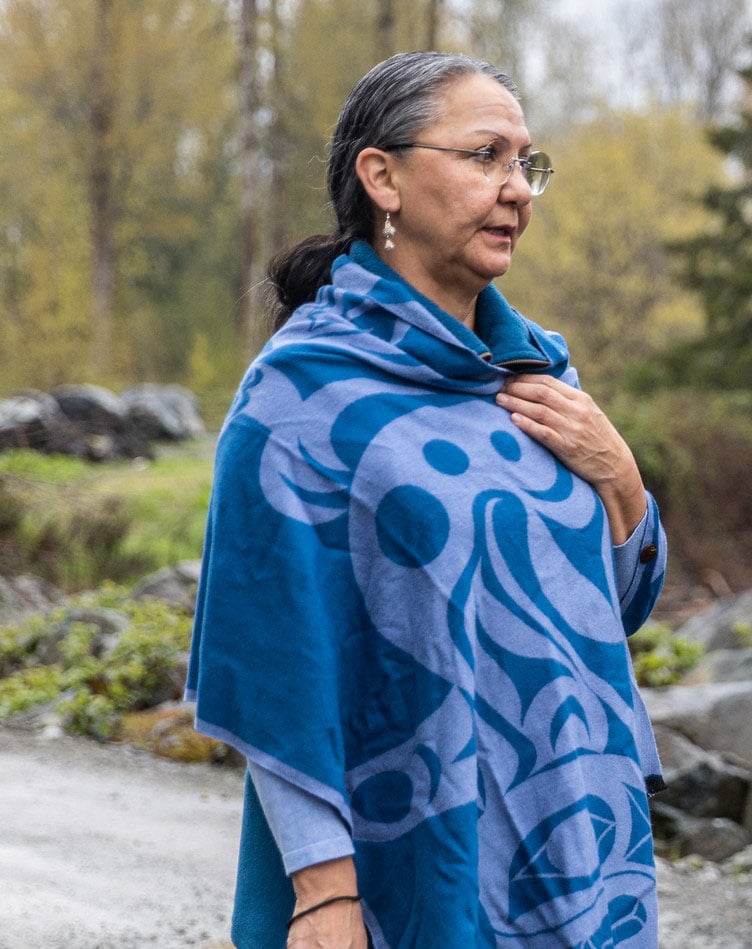 Lydia Hwitsum stands in a forest wrapped in a blue shawl with a First Nations design. She holds her hand to her heart and looks on.