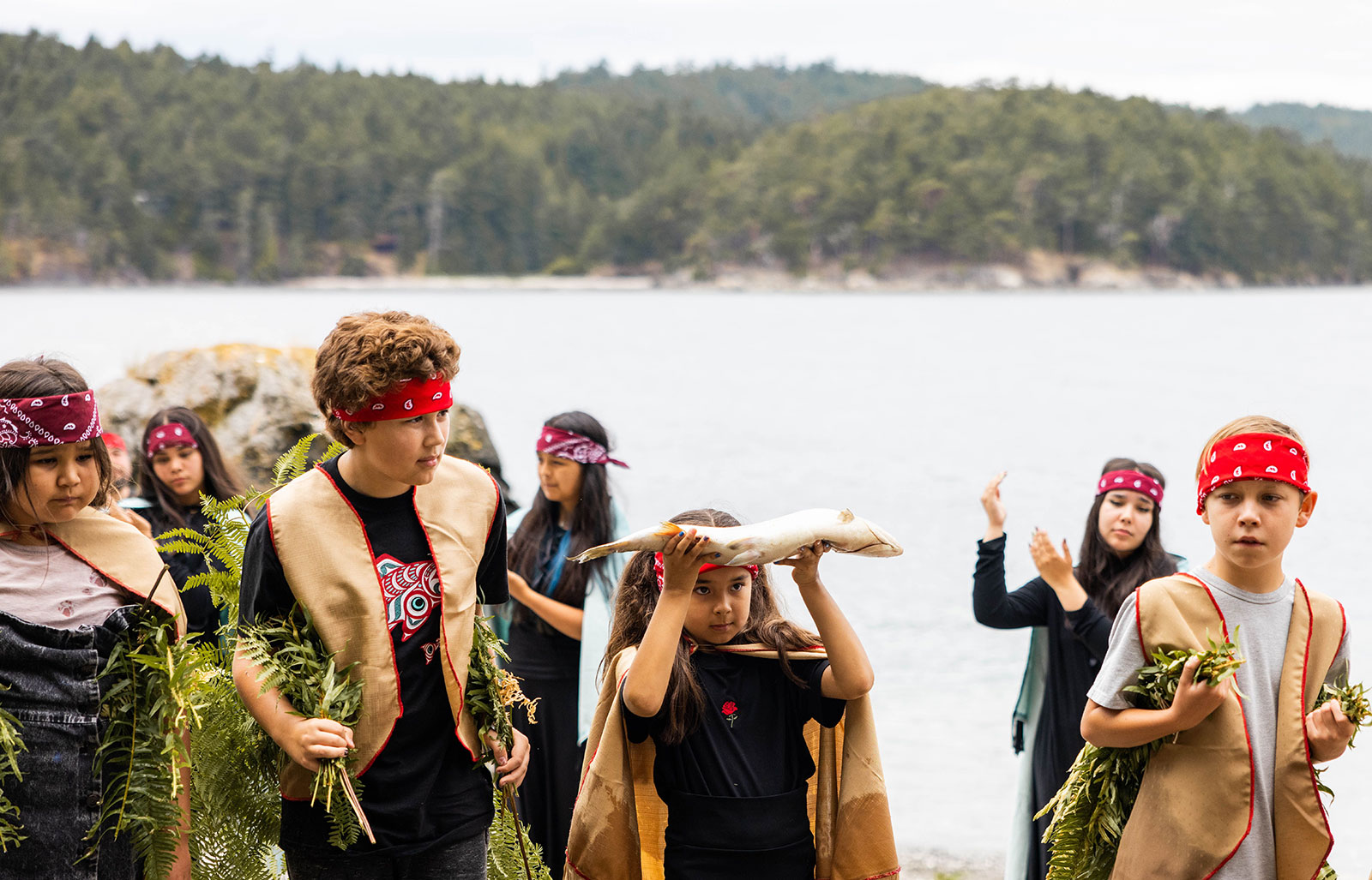 A group of young children stand on a beach. Folded red scarves are tied around their heads. Most are holding ferns. One, at centre, is holding a fish above her head.