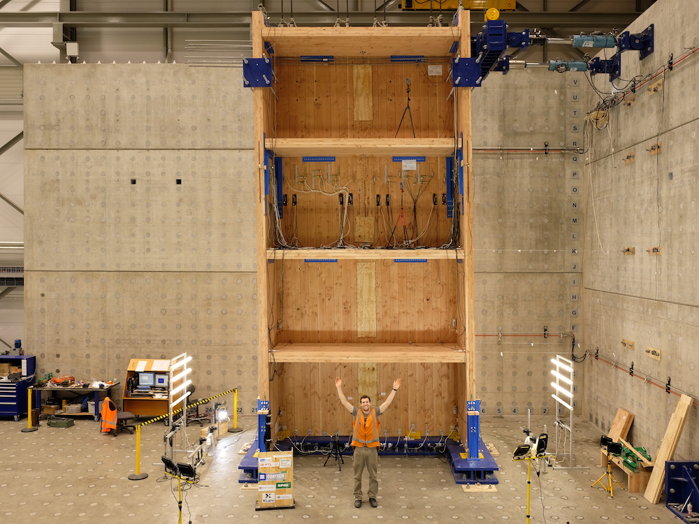A man in a hard hat and safety vest stands in front of a four-storey frame made out of mass timber panels.