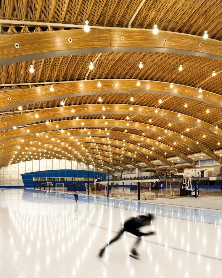 A skater on ice in an arena under an arching wooden roof with many components.
