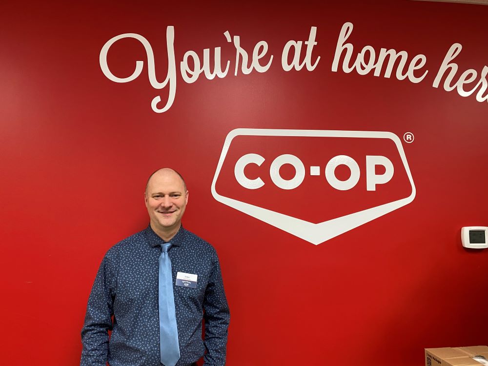 A light-skinned man wearing a patterned dark blue shirt and a light blue tie stands in front of a red wall with the words 'You're at home here' and 'CO-OP' painted in white.