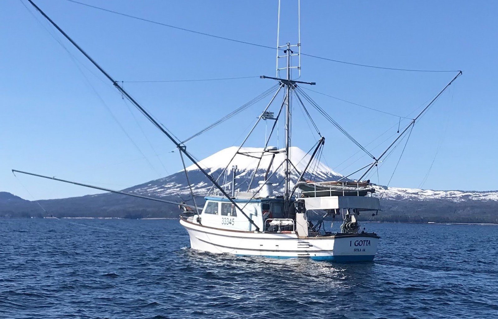 A fishing boat with the name 'I Gotta' painted on its stern trolls the waters with a snow-capped volcanic peak in the background.