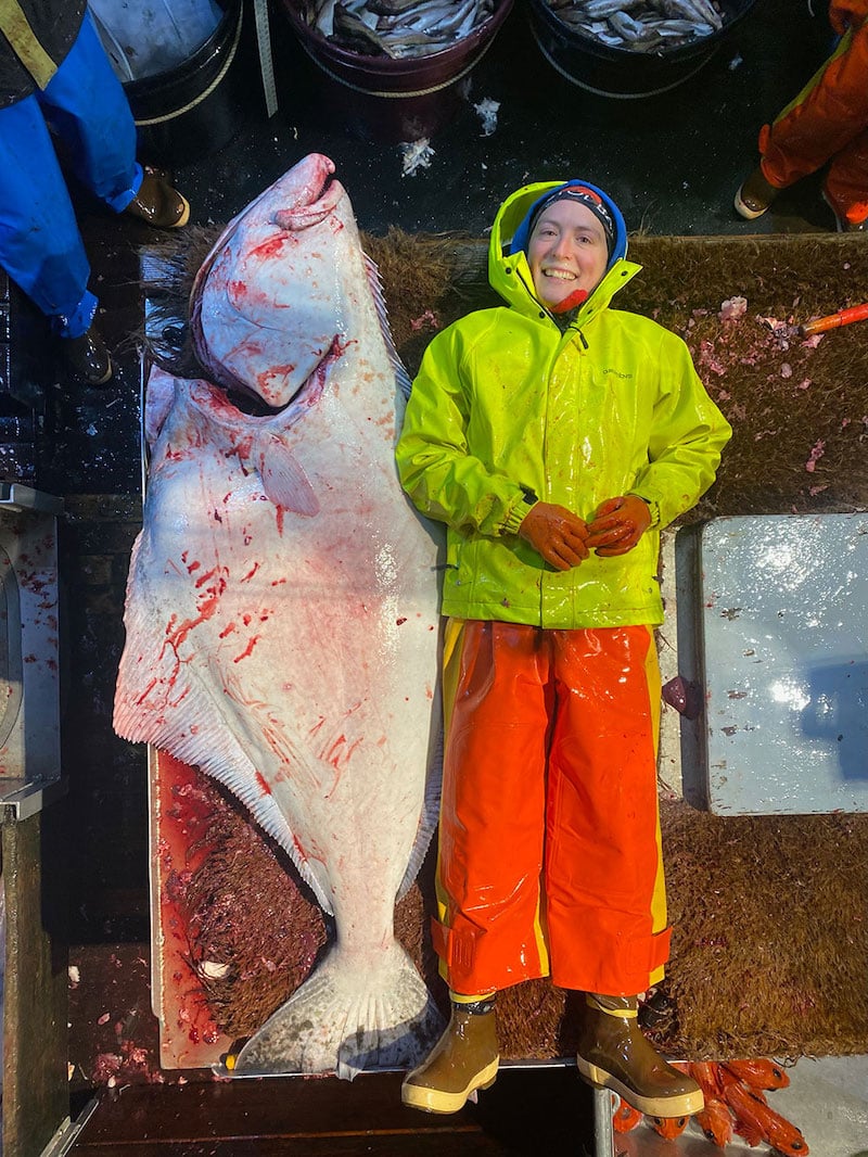 A woman with light skin wearing neon green and orange rain gear lies next to a landed halibut that is slightly larger than she is.
